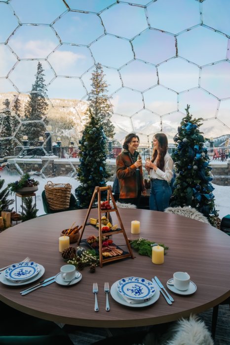 A happy couple stands in a geodome outdoors surrounded by mountains smiling as they each have a glass of champagne with a table beautifully set out for two.