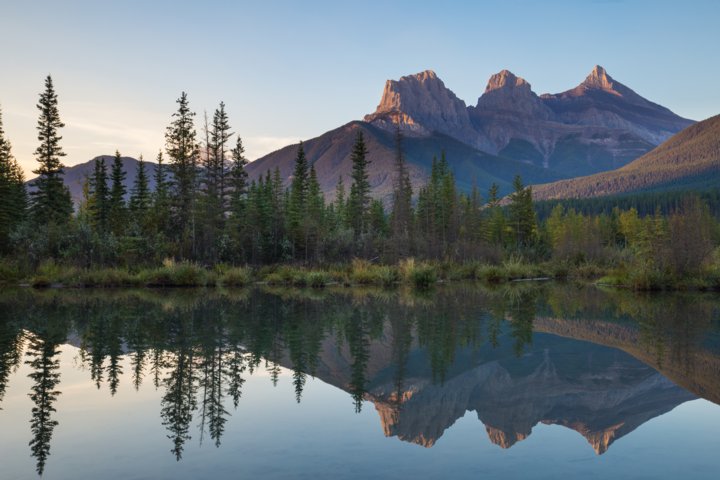 Sunrise view of the Three Sisters peaks in Canmore with their reflection in a pond.