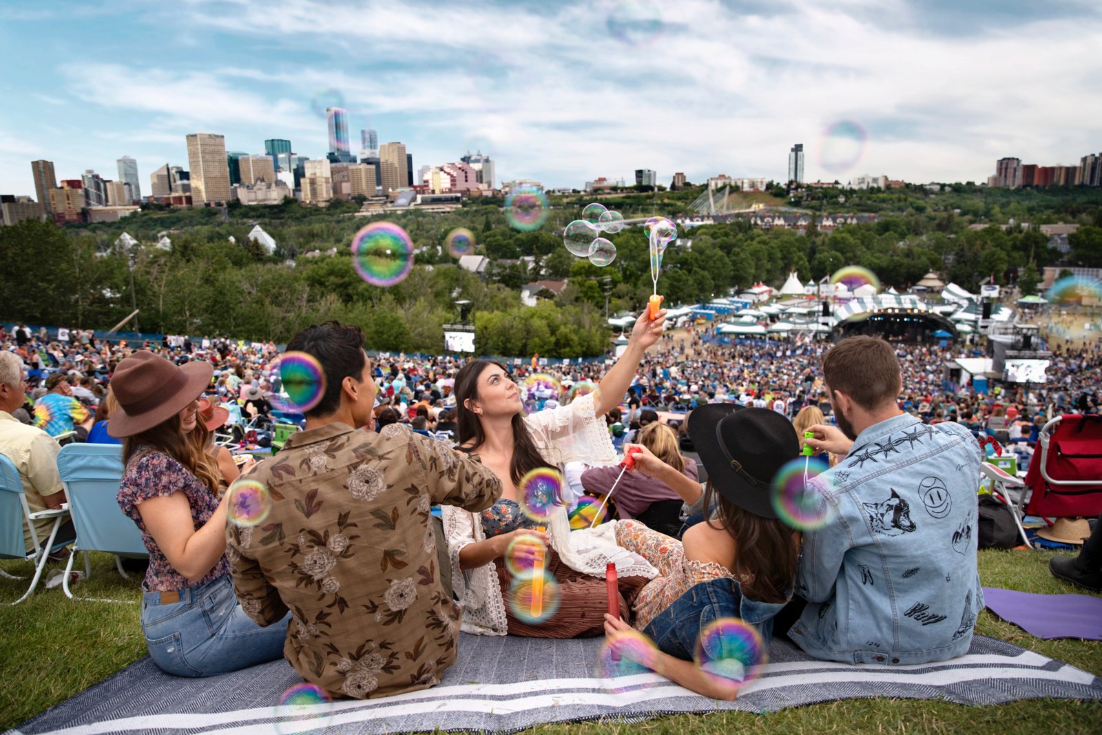 A group of people sit on a hill overlooking the city skyline at the Edmonton Folk Music Festival.