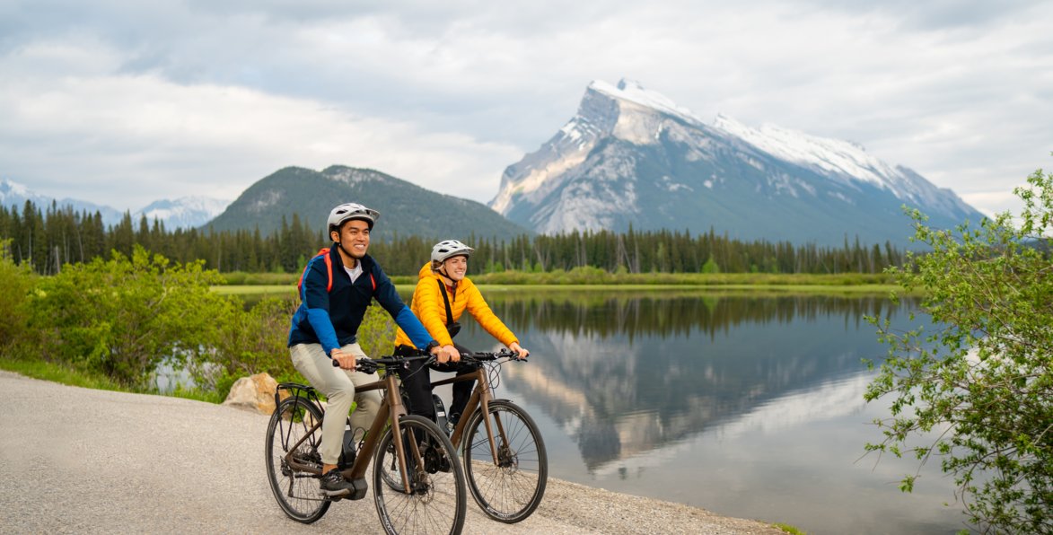 A couple rides bikes on a path with a mountain and lake in the background.
