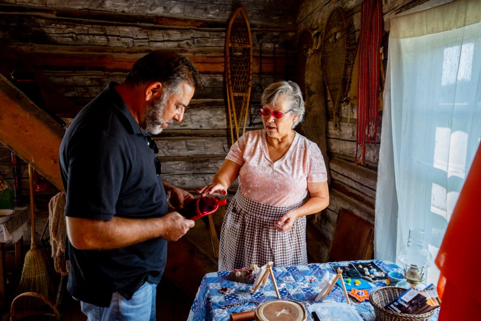 An Indigenous woman instructs a man holding a fabric craft inside a historic cabin.