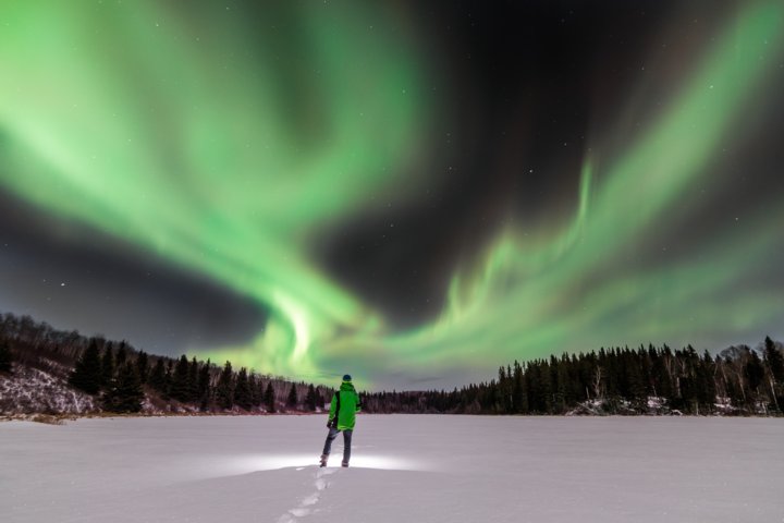 Someone standing in the middle of a snow covered frozen lake watching the night sky light up with colour from the Northern Lights