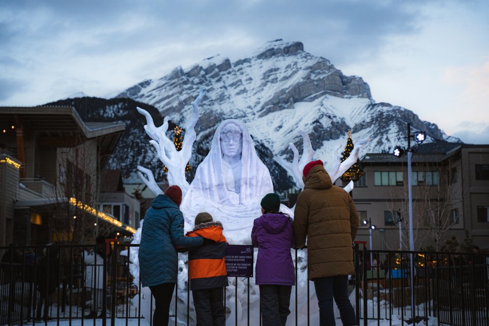 A family looking at a snow sculpture with mountains in the background.