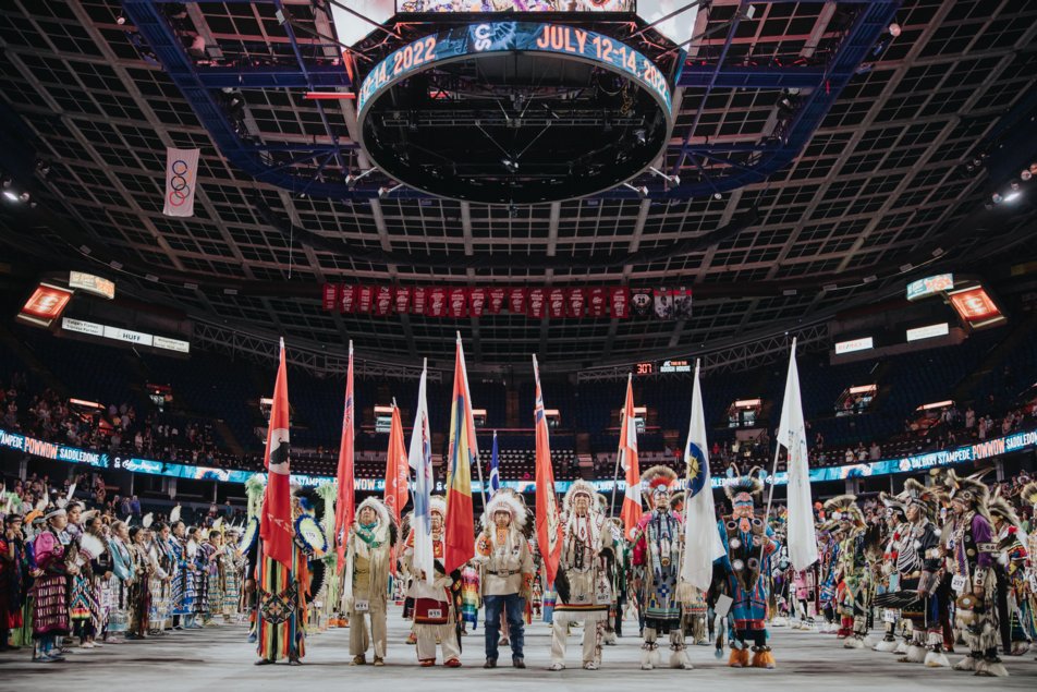 Members of the Indigenous community dressed in regalia hold flags to kick off an event at Saddledome as the crowd looks on.
