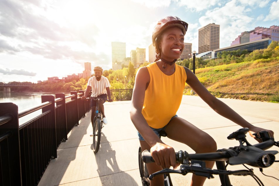 A man and a woman bike along a path beside the river with Edmonton’s downtown in the background.