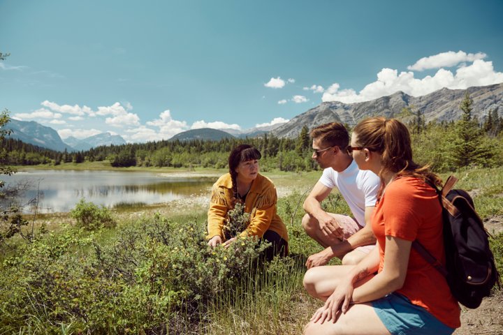 A woman in traditional Indigenous clothing shows some plants to a young man and woman kneeling beside a lake in the mountains.