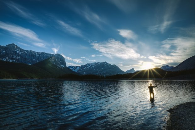 Scenic shot of man fly fishing in a lake surrounded by mountains at sunset in Kananaskis Country.