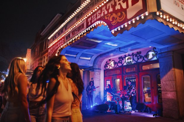 Group of girls walking by a the front of a theatre at night with lit signage.