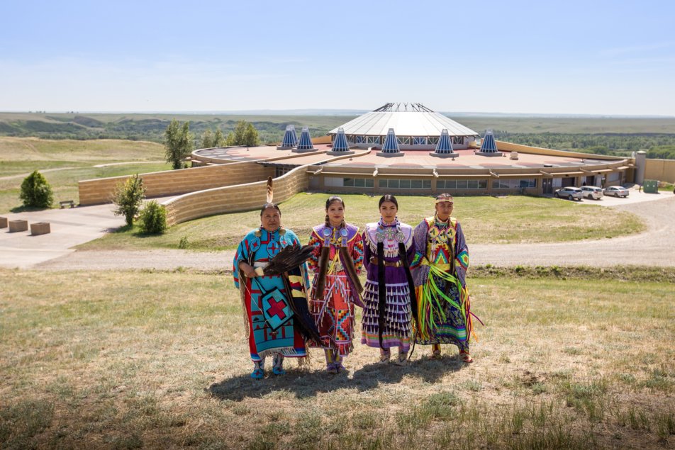 Four women in traditional Indigenous clothing stand in front of the visitor centre at Blackfoot Crossing Historical Park.