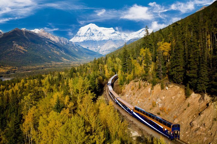 A passenger train passing through a forest with a mountain view in the background.