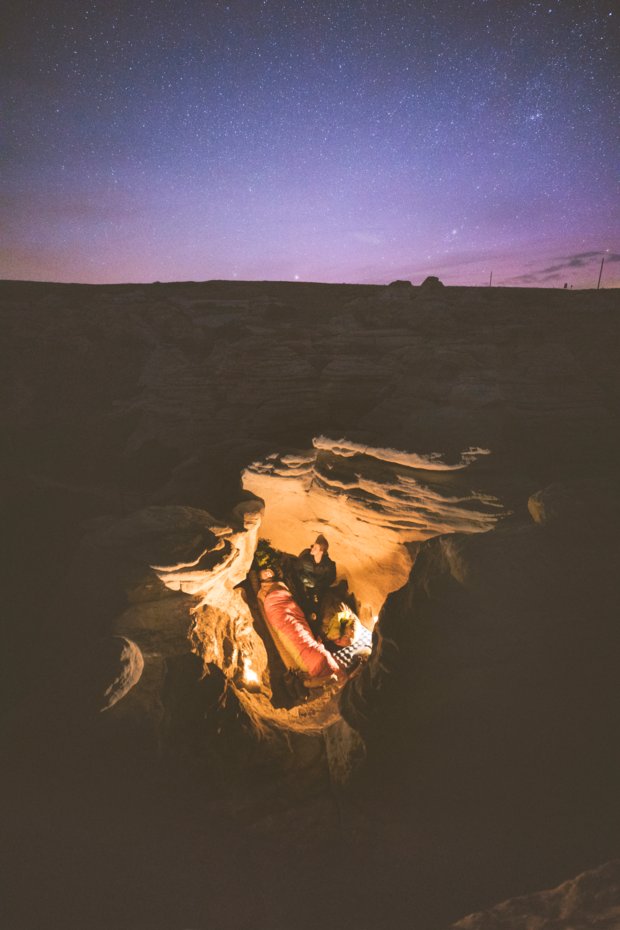 A man and woman camping inside hoodoos while a fire burns next to them and the stars light up the nights sky.