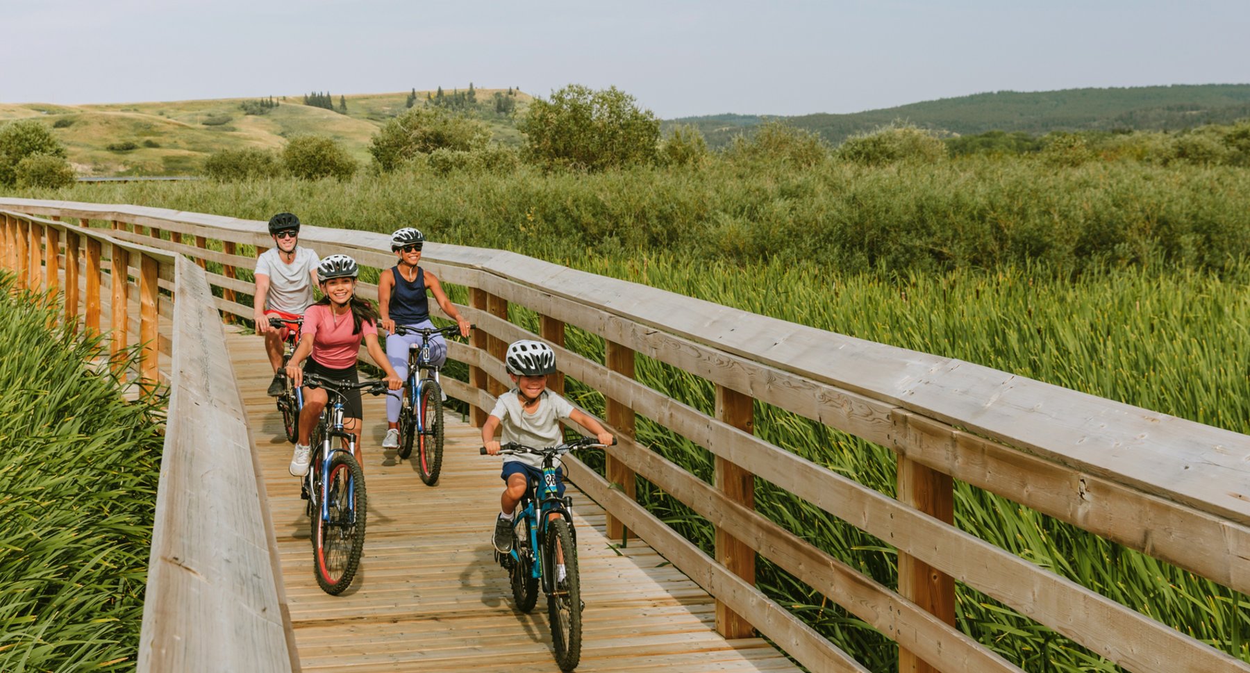Family of four smiling as they bike along a boardwalk over tall grass.