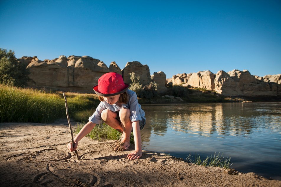 young girl draws in the sand in with a stick beside the Milk River in Cypress Hills Provincial Park