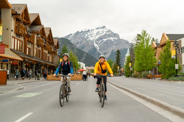 Two friends riding their bikes down Banff Avenue with the mountains in the background.