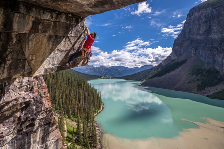 Man rock climbing the side of a mountain above a lake with a hotel in the distant background