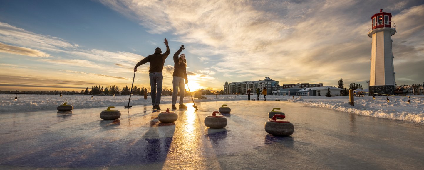 People curling on a frozen lake near a lighthouse in the setting sun.