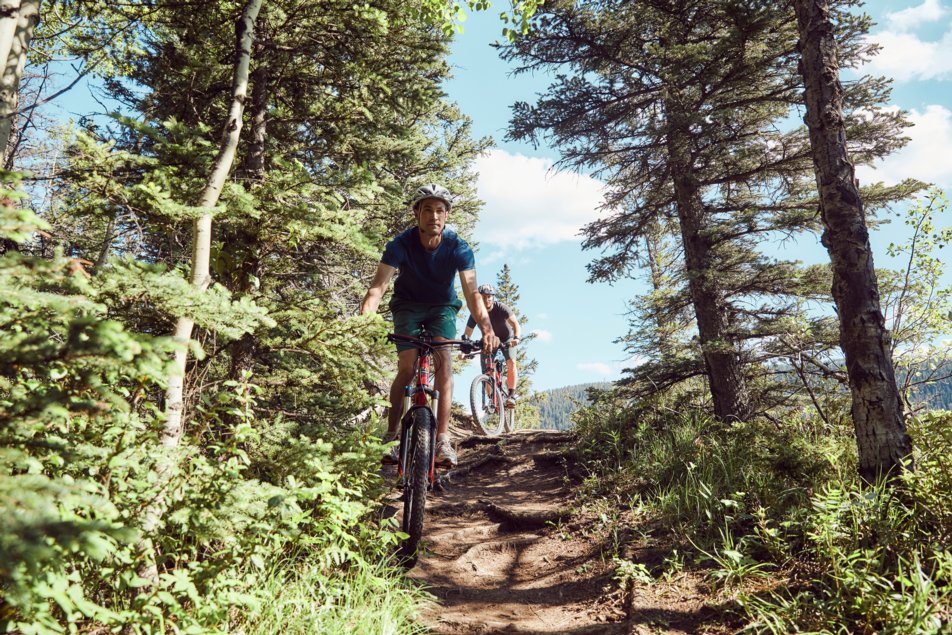 Two men ride mountain bikes down a rutted path in the forest.