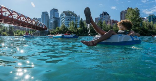 oung friends in swimsuits & buoyancy aids float leisurely on inner tubes down the Bow River in Calgary. Tree-lined banks & distant cityscape provide scenic backdrop as they approach Peace Bridge on sunny day, creating a peaceful & carefree atmosphere.