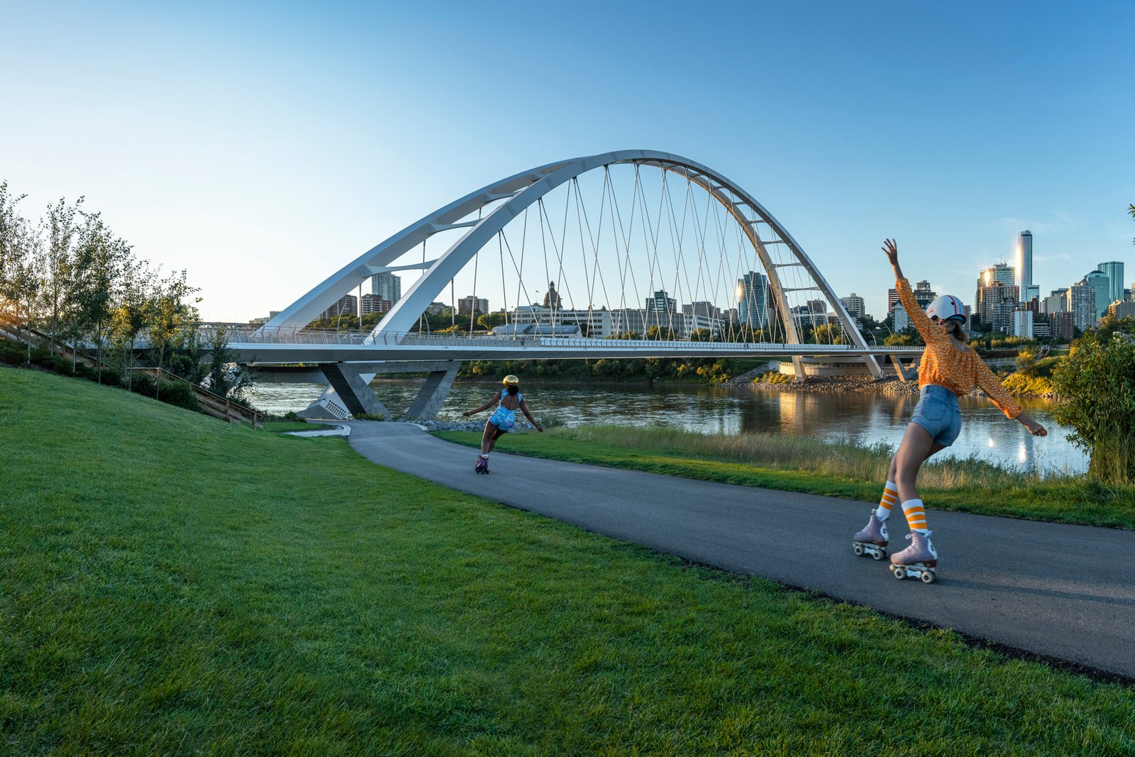 Two young women in summer clothes roller skate on pathway by North Saskatchewan River in Edmonton, heading towards Walterdale Bridge. City skyline shimmers in background, creating a fun atmosphere.
