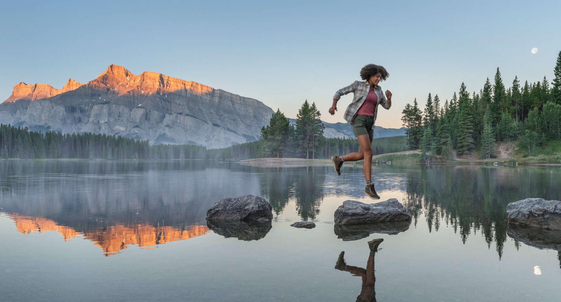 Woman jumping from rock to rock at on a lake with a mountain in the background.