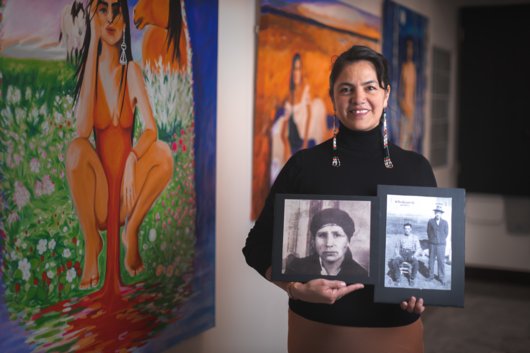 Indigenous artist Lana Whiskeyjack stands in front of her original artwork holding photos of her family members.